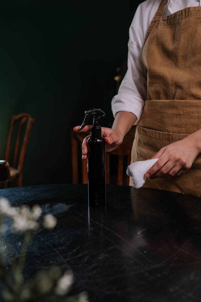 A waitress in an apron cleaning a table with a spray bottle and cloth in a dimly lit restaurant.